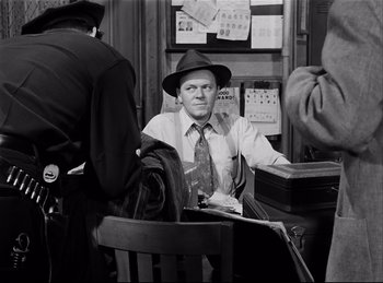 Movie still from “Detective Story” (1951), directed by William Wyler – A man sitting at a table wearing a hat and a tie; Medium shot, Over the shoulder angle