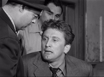 Movie still from “Detective Story” (1951), directed by William Wyler – A man in a suit and tie talking to two other men; Medium shot, Over the shoulder angle