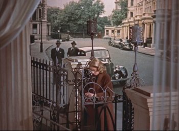 Movie still from “Dial M for Murder” (1954), directed by Alfred Hitchcock – A woman standing on the side of a road next to a fence; Wide shot, High angle