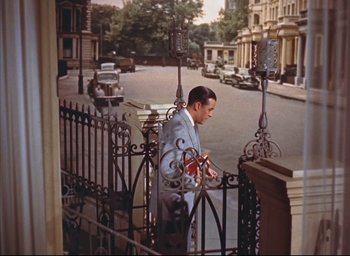 Movie still from “Dial M for Murder” (1954), directed by Alfred Hitchcock – A man standing on a porch holding a guitar; Wide shot, High angle