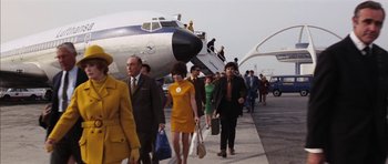 Movie still from “Diamonds Are Forever” (1971), directed by Guy Hamilton – A group of people walking down a sidewalk next to an airplane; Wide shot, Low angle