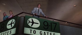 Movie still from “Diamonds Are Forever” (1971), directed by Guy Hamilton – A man in a suit and tie standing next to an airport sign; Wide shot, Low angle