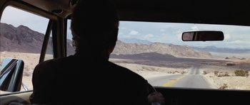 Movie still from “Diamonds Are Forever” (1971), directed by Guy Hamilton – A person driving a car on the side of a road; Wide shot, Over the shoulder angle