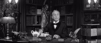 Movie still from “Diary of a Chambermaid” (1964), directed by Luis Buñuel – An older man sitting at a desk in front of a book case; Medium shot, Low angle