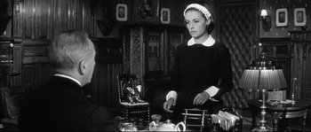 Movie still from “Diary of a Chambermaid” (1964), directed by Luis Buñuel – A woman in a maid's outfit standing in front of a chair; Medium shot, Over the shoulder angle