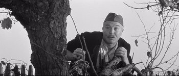 Movie still from “Diary of a Chambermaid” (1964), directed by Luis Buñuel – A man leaning up against a tree looking at the camera; Medium shot, Low angle
