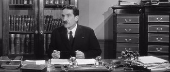 Movie still from “Diary of a Chambermaid” (1964), directed by Luis Buñuel – A man sitting at a desk in a suit and tie; Medium shot, Low angle