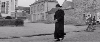 Movie still from “Diary of a Chambermaid” (1964), directed by Luis Buñuel – A man walking down the street wearing a long coat and a hat; Wide shot, Low angle