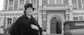 Movie still from “Diary of a Chambermaid” (1964), directed by Luis Buñuel – An older woman wearing a hat and holding a purse in front of a building; Medium shot, Low angle