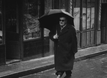 Movie still from “Diary of a Pregnant Woman” (1958), directed by Agnès Varda – An older man walking down the street holding an umbrella; Wide shot, High angle