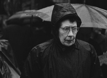 Movie still from “Diary of a Pregnant Woman” (1958), directed by Agnès Varda – An older woman wearing a raincoat and holding an umbrella; Close Up shot, Over the shoulder angle