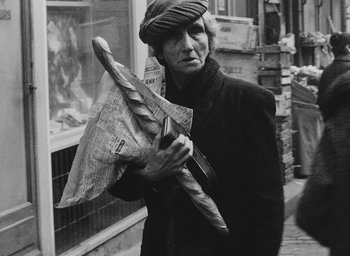Movie still from “Diary of a Pregnant Woman” (1958), directed by Agnès Varda – An older woman holding a newspaper umbrella in the street; Medium shot, Over the shoulder angle