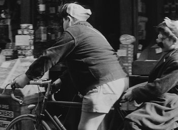 Movie still from “Diary of a Pregnant Woman” (1958), directed by Agnès Varda – A man riding a bike down a street; Medium shot, Over the shoulder angle