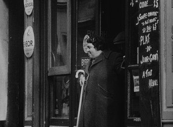 Movie still from “Diary of a Pregnant Woman” (1958), directed by Agnès Varda – An older woman holding a cane in front of a store; Medium shot, High angle