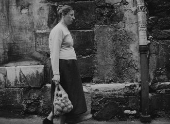 Movie still from “Diary of a Pregnant Woman” (1958), directed by Agnès Varda – A black and white photo of a woman carrying a bag of fruit; Wide shot, Low angle