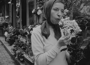 Movie still from “Diary of a Pregnant Woman” (1958), directed by Agnès Varda – A young woman wearing a head scarf is blowing on some flowers; Close Up shot, Low angle