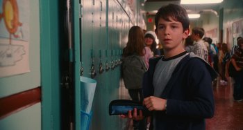 Movie still from “Diary of a Wimpy Kid” (2010), directed by Thor Freudenthal – A young man holding a cell phone in a school hallway; Medium shot, Over the shoulder angle