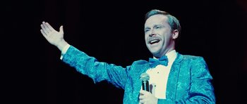 Movie still from “Diary of a Wimpy Kid: Rodrick Rules” (2011), directed by David Bowers – A man in a bow tie holding a microphone and smiling; Medium shot, Low angle