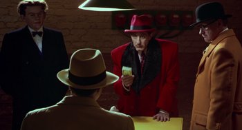 Movie still from “Dick Tracy” (1990), directed by Warren Beatty – A man in a red coat and a hat is holding a card; Medium shot, Over the shoulder angle