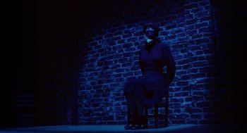 Movie still from “Dick Tracy” (1990), directed by Warren Beatty – A woman sitting on a chair in front of a brick wall; Wide shot, High angle