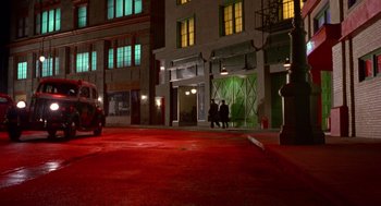 Movie still from “Dick Tracy” (1990), directed by Warren Beatty – Three people are sitting on the sidewalk in front of a building at night; Extreme Wide shot, High angle