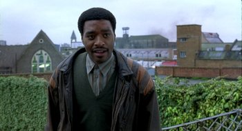 Movie still from “Dirty Pretty Things” (2002), directed by Stephen Frears – A man standing in front of a building with trees in the background; Medium shot, Over the shoulder angle