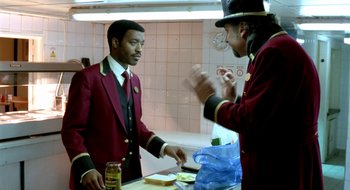 Movie still from “Dirty Pretty Things” (2002), directed by Stephen Frears – A man in a red jacket is cutting a sandwich; Medium shot, Over the shoulder angle