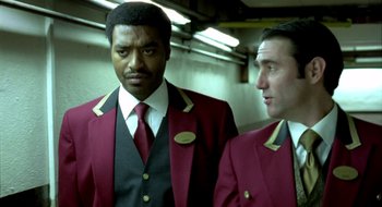 Movie still from “Dirty Pretty Things” (2002), directed by Stephen Frears – A man in a suit and tie standing next to another man in a suit and tie; Medium shot, Low angle