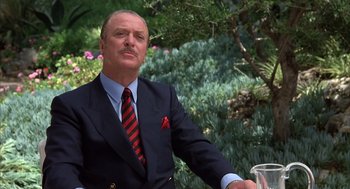 Movie still from “Dirty Rotten Scoundrels” (1988), directed by Frank Oz – A man in a suit and tie sitting in front of some bushes; Close Up shot, Low angle