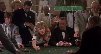 Movie still from “Dirty Rotten Scoundrels” (1988), directed by Frank Oz – A group of people standing around a table with cards; Medium shot, High angle