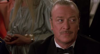 Movie still from “Dirty Rotten Scoundrels” (1988), directed by Frank Oz – An older man in a tuxedo sitting in front of a group of people; Close Up shot, Over the shoulder angle