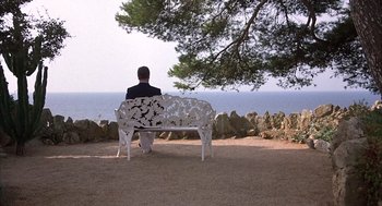 Movie still from “Dirty Rotten Scoundrels” (1988), directed by Frank Oz – A man sitting on top of a white bench near the ocean; Wide shot, High angle