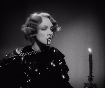Movie still from “Dishonored” (1931), directed by Josef von Sternberg – A black and white photo of a woman smoking a cigarette next to a lit candle; Medium shot, Low angle