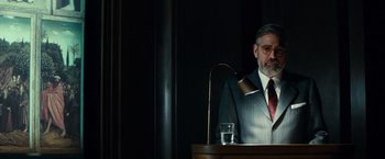 Movie still from “The Monuments Men” (2014), directed by George Clooney – A man standing at a podium in front of a glass of water; Medium shot, Over the shoulder angle