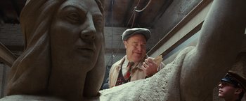 Movie still from “The Monuments Men” (2014), directed by George Clooney – A man in a hat and tie working on a statue; Medium shot, Over the shoulder angle