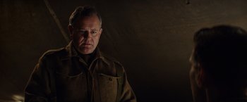 Movie still from “The Monuments Men” (2014), directed by George Clooney – An older man wearing a brown jacket and standing in front of a dark background; Close Up shot, Low angle