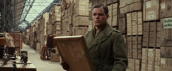 Movie still from “The Monuments Men” (2014), directed by George Clooney – A man in a trench coat holding a wooden box; Medium shot, Over the shoulder angle