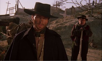 Movie still from “Django” (1966), directed by Sergio Corbucci – A man in a hat and a black coat standing in front of a cemetery; Medium shot, Low angle