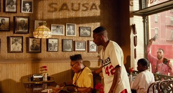 Movie still from “Do the Right Thing” (1989), directed by Spike Lee – Two men are standing in front of a wall with pictures on it; Medium shot, Over the shoulder angle