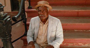 Movie still from “Do the Right Thing” (1989), directed by Spike Lee – An older man sitting on a set of stairs wearing a hat; Medium shot, High angle