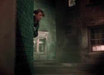 Movie still from “Doctor X” (1932), directed by Michael Curtiz – A man wearing a hat leaning up against a brick wall; Medium shot, High angle
