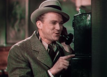 Movie still from “Doctor X” (1932), directed by Michael Curtiz – A man wearing a suit and a hat talking on a phone; Medium shot, Low angle