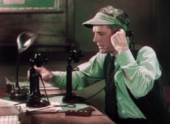 Movie still from “Doctor X” (1932), directed by Michael Curtiz – An older man wearing a green shirt and a hat is sitting at a table with a phone; Medium shot, High angle