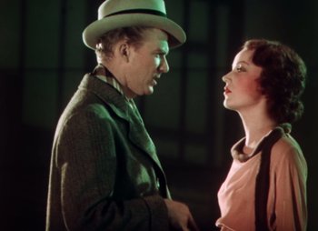 Movie still from “Doctor X” (1932), directed by Michael Curtiz – An older man wearing a hat standing next to an older woman; Medium shot, Over the shoulder angle