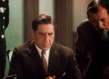 Movie still from “Doctor X” (1932), directed by Michael Curtiz – A man wearing a suit and tie sitting at a table; Medium shot, Over the shoulder angle
