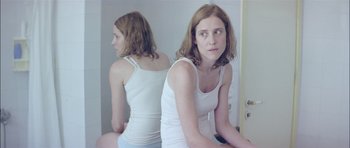 Movie still from “Dogtooth” (2009), directed by Yorgos Lanthimos – A woman in a white tank top looking at her reflection in a mirror; Medium shot, Over the shoulder angle