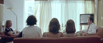 Movie still from “Dogtooth” (2009), directed by Yorgos Lanthimos – Three people sitting on a couch looking out a window; Medium shot, Over the shoulder angle