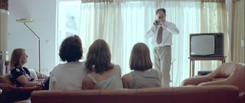 Movie still from “Dogtooth” (2009), directed by Yorgos Lanthimos – A group of people sitting on a couch and a man taking a picture; Medium shot, Low angle