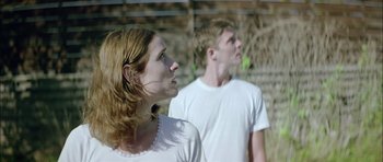 Movie still from “Dogtooth” (2009), directed by Yorgos Lanthimos – A woman and a man standing next to each other in front of a fence; Medium shot, Low angle