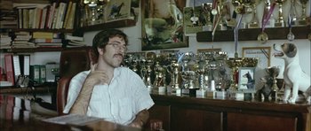 Movie still from “Dogtooth” (2009), directed by Yorgos Lanthimos – A man sitting in front of many trophies; Medium shot, Low angle